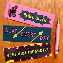 Load image into Gallery viewer, The other three bookmarks pictured here with the pink pen. One has her riding a motorbike with her cape flapping in the wind, above the caption “rebel queen”; another with the caption “slay every day” in between spears; and the last with the caption “veni.vidi.incanvidi” (Latin for “I came, I saw, I set it ablaze”) surrounded by fire.