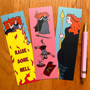 Three bookmarks laid out each with a different image celebrating Boudicca. One shows her wielding a spear with a crown in her ginger hair; another raising it above a blaze of fire captioned “raise some hell”; another having dispatched a Roman soldier who is tumbling down the bookmark with his weapon and armour falling around him. Pictured here with a pink pen to match the pink background of some of the bookmarks.