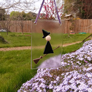 Photo of glasswork which depicts a witch in a minimalist style, with a black dress and black hat, flying on a broomstick. The artwork hands from a purple thread outside, with a fence and grass and flowers in the background.