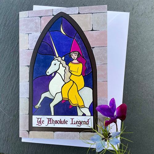 Photo of greetings card depicting a stained glass window set in a stone wall. The glass design shows a woman in mediaeval dress with a pointed hat riding a unicorn against a moonlit night sky. The caption reads 'Ye Absolute Legend'. The card is with a white envelope and two blue/purple flowers on a slate background.