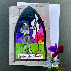 Photo of greetings card depicting a stained glass window set in a stone wall. The glass design shows a woman in mediaeval armour with chain maille, bearing a white flag and a sword, against a sunrise sky. The caption reads 'You've Got Maille'. The card is with a white envelope and two blue/purple flowers on a slate background.