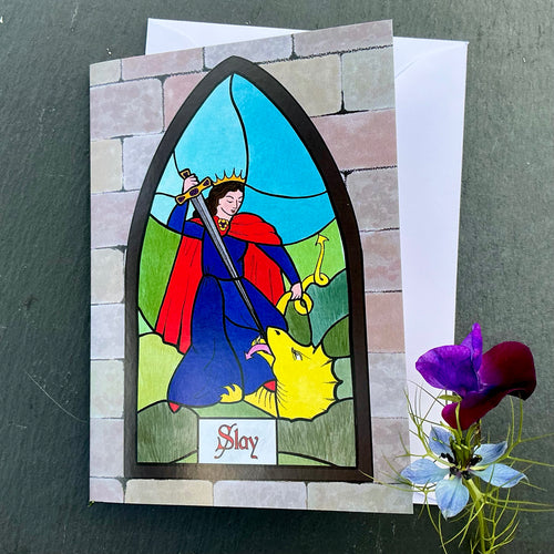 Photo of greetings card depicting a stained glass window set in a stone wall. The glass design shows a woman in mediaeval blue dress in a red cape and crown, slaying a dragon with her sword. The caption reads 'Slay'. The card is with a white envelope and two blue/purple flowers on a slate background.