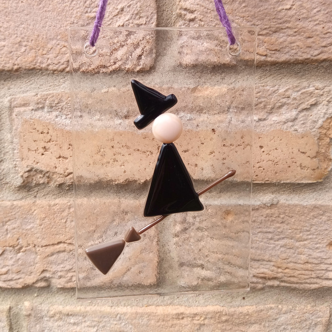 Photo of glasswork which depicts a witch in a minimalist style, with a black dress and black hat, flying on a broomstick. The artwork hands from a purple thread against a brick wall.