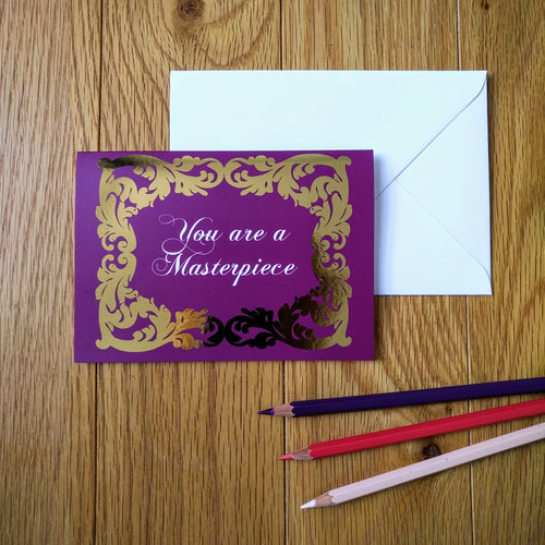 Photo of a greeting card in a purple colour with ornate gold leaf styling around the edges. In a white baroque font at the centre is the phrase “You are a Masterpiece”. Next to the card is a white envelope and three coloured pencils.