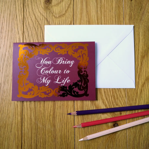 Photo of a greeting card in a crimson colour with ornate gold leaf styling around the edges. In a white baroque font at the centre is the phrase “You Bring Colour to My Life”. Next to the card is a white envelope and three coloured pencils.