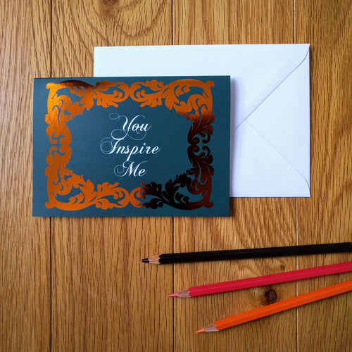 Photo of a greeting card in a dark blue-green colour with ornate gold leaf styling around the edges. In a white baroque font at the centre is the phrase “You Inspire Me”. Next to the card is a white envelope and three coloured pencils.