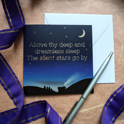 Photo of a card that shows a dark night. We see the silhouette of a valley, a row of trees, and a house from which we see smoke coming from the chimney. The sky is full of stars and a waning moon. Written in the sky in gold foil are the words “Above thy deep and dreamless sheep The silent stars go by”. Next to the card is a white envelope, silver pen, and a curling purple ribbon.